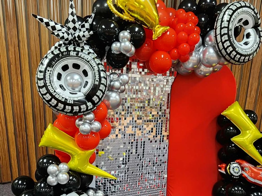 Racing-themed balloon installation in black, red and silver with checkered accents, lightning-bolt foils and tire balloons against a sequin panel